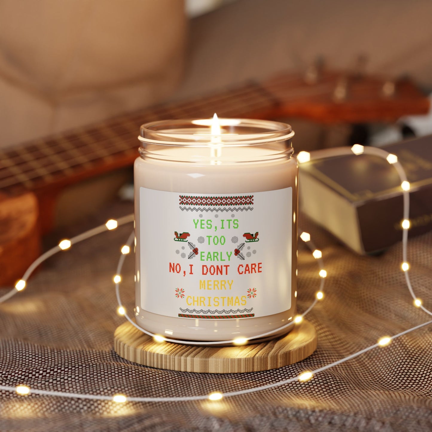 Candle with a humorous Christmas label on a wooden coaster surrounded by string lights and a guitar in the background.