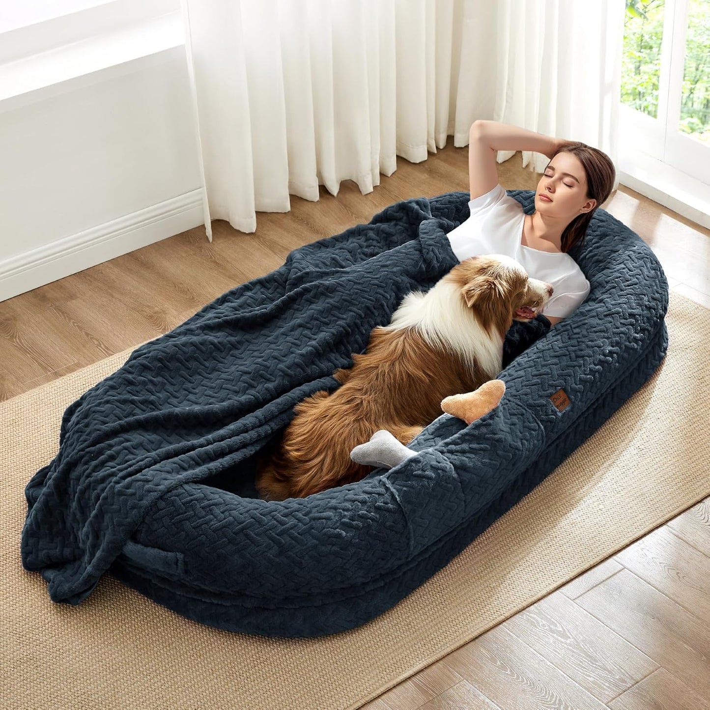 Woman and dog lying in a large blue pet bed in a bright room with white curtains.