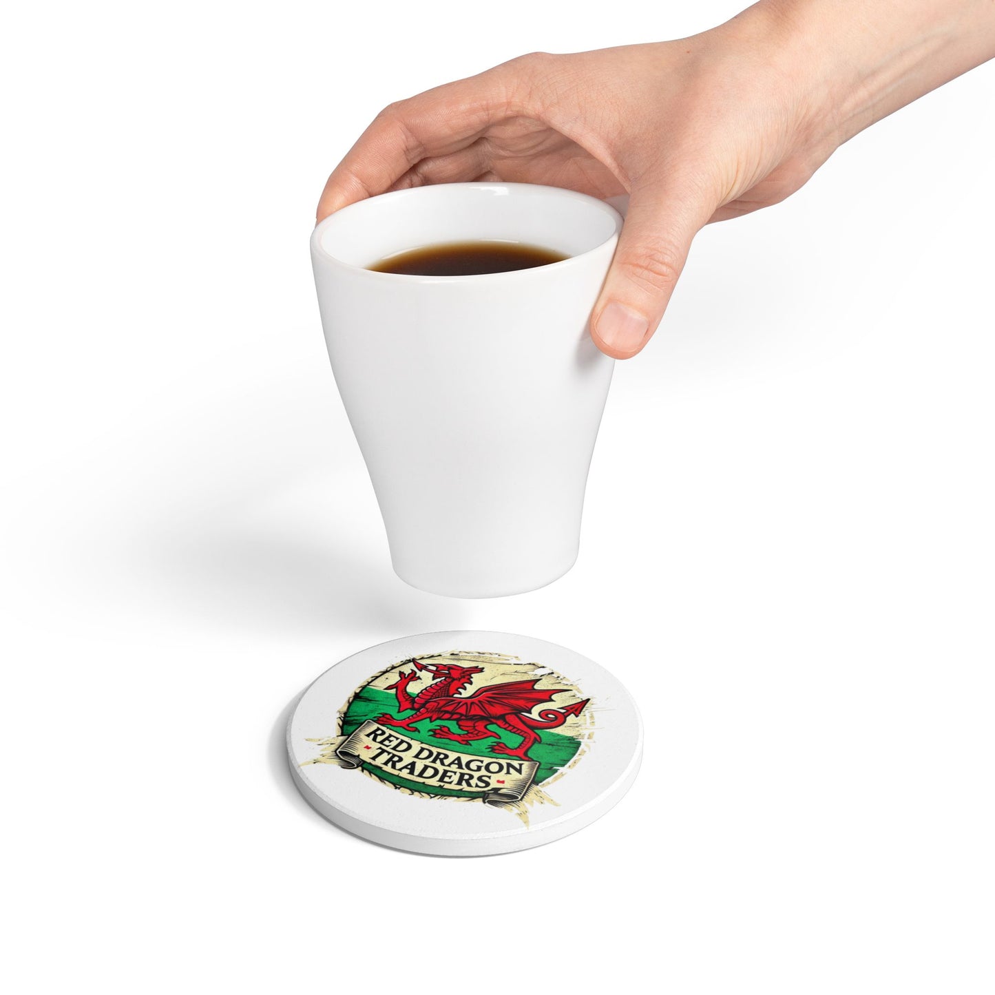 Hand holding a white mug with a Red Dragon Traders coaster underneath on a white background