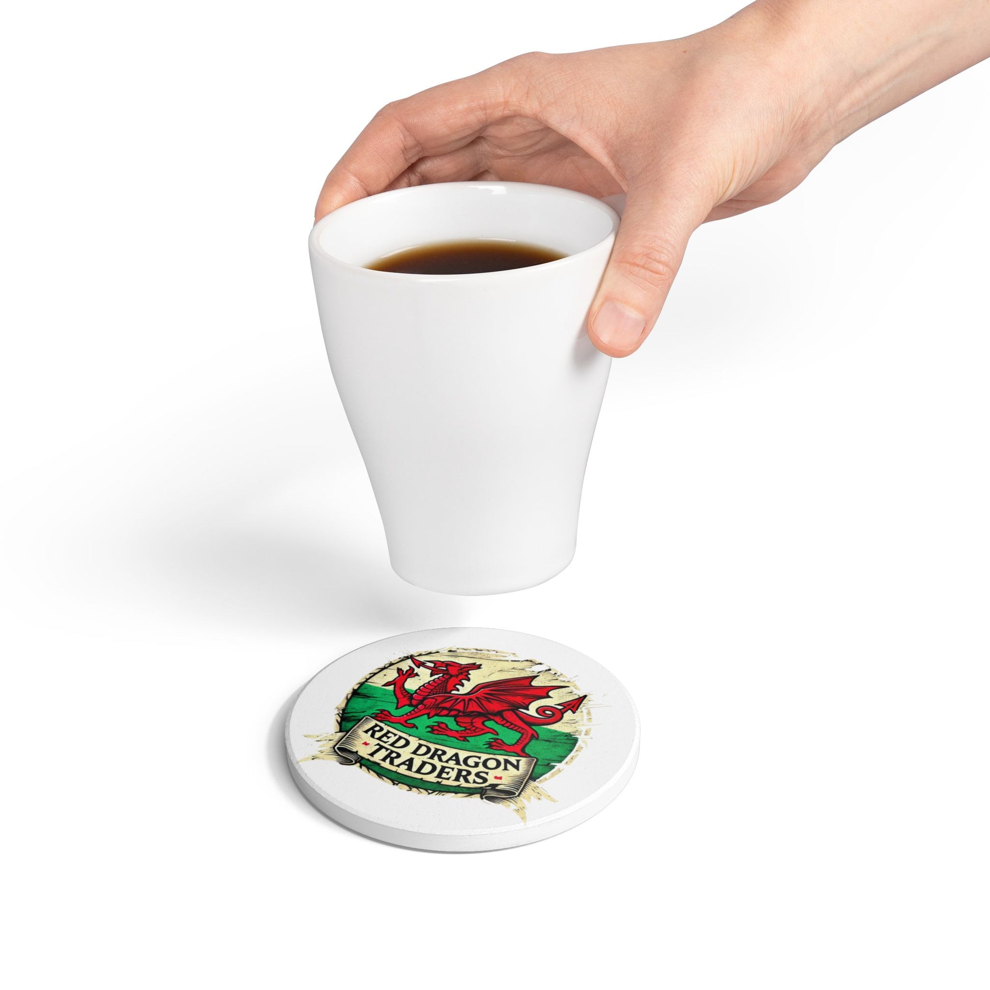 Hand holding a white mug with a Red Dragon Traders coaster underneath on a white background