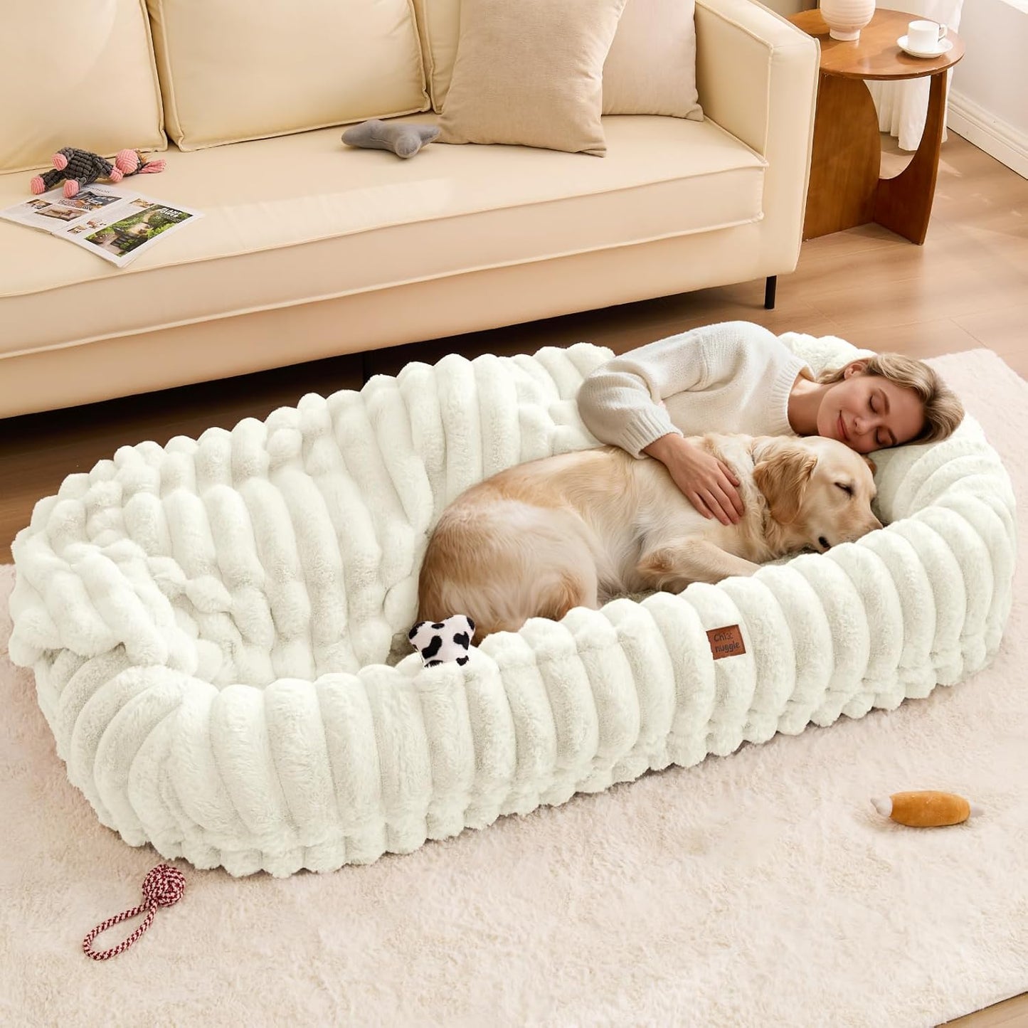 Woman lying on a large white pet bed with a dog, in a living room setting.