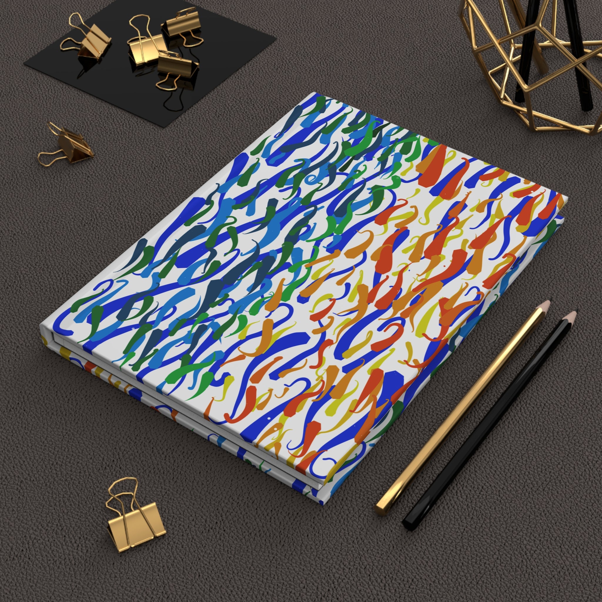 Hardcover journal with a colorful abstract squiggle pattern, shown on a grey desk with pencils and binder clips.