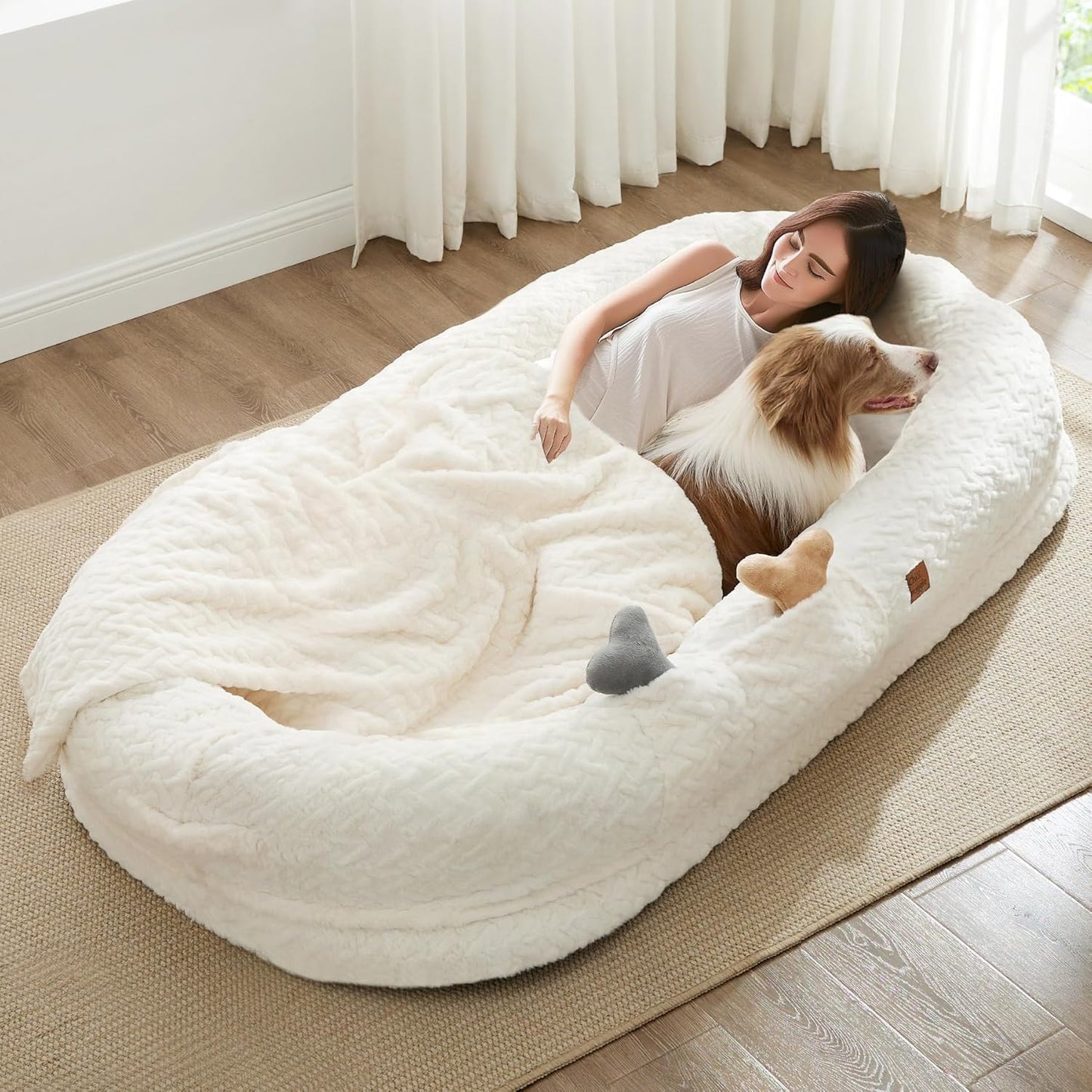 Person lying on a large white pet bed with a dog inside, in a bright room with curtains.