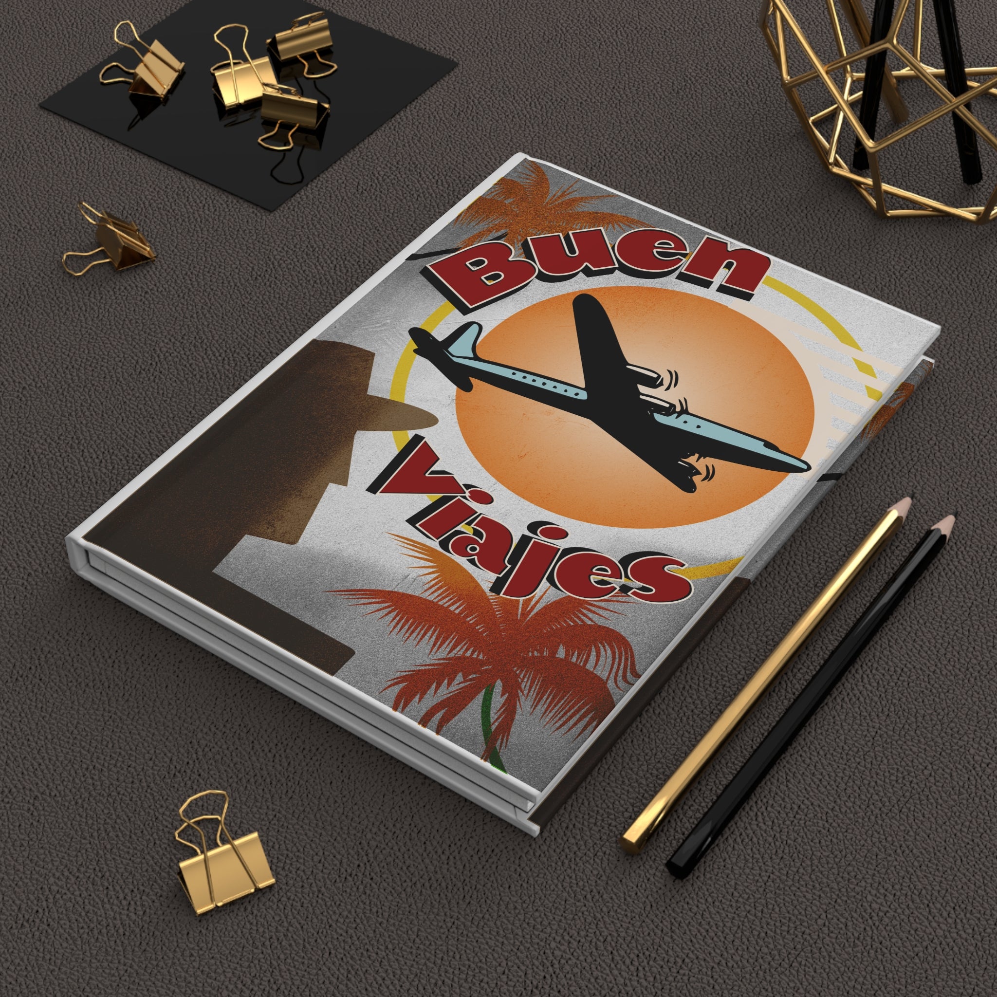 Hardcover journal with a retro 'Buen Viajes' airplane and sunset travel design, styled on a desk