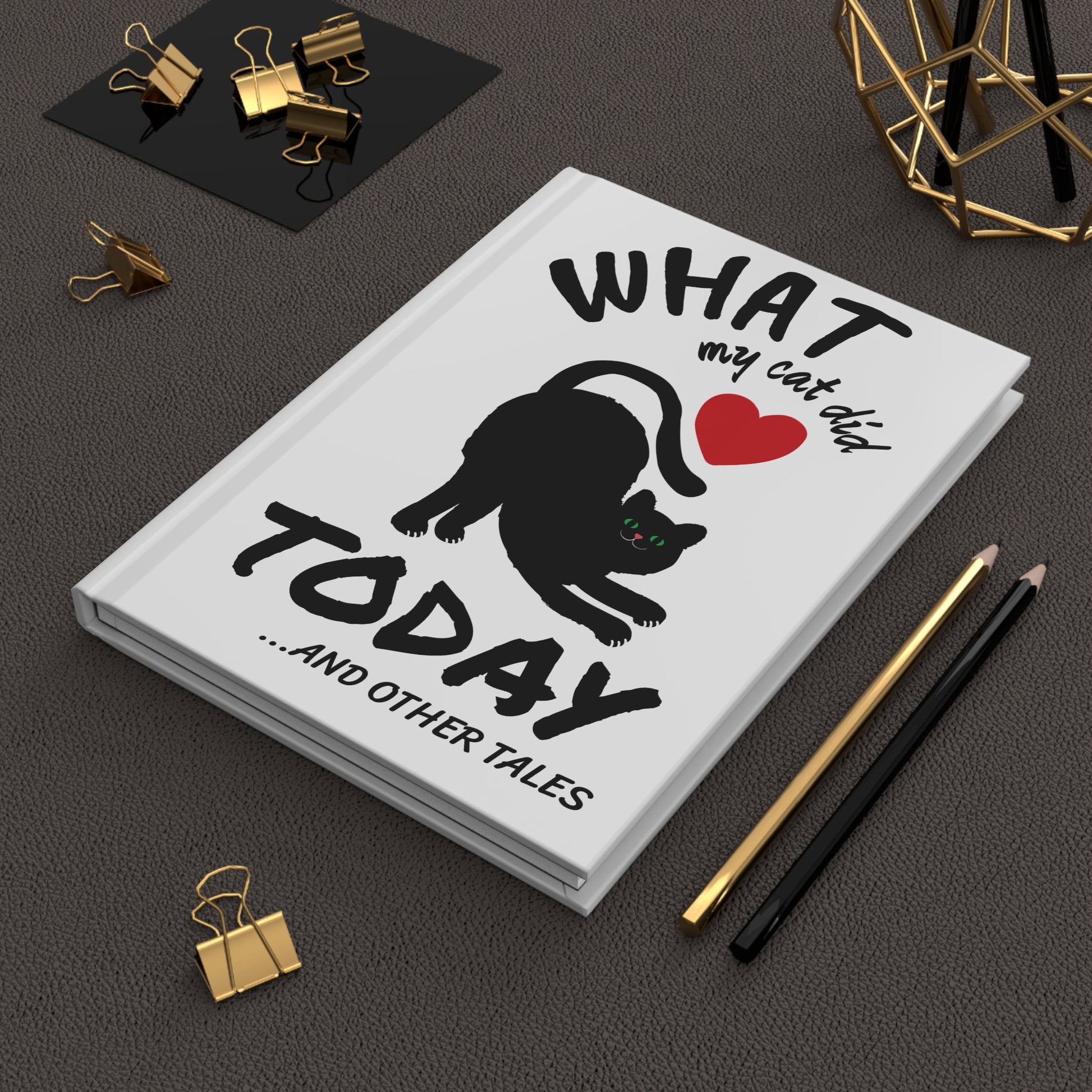 Cat Journal titled 'What my cat did today...and other tales' on a dark surface with office supplies.