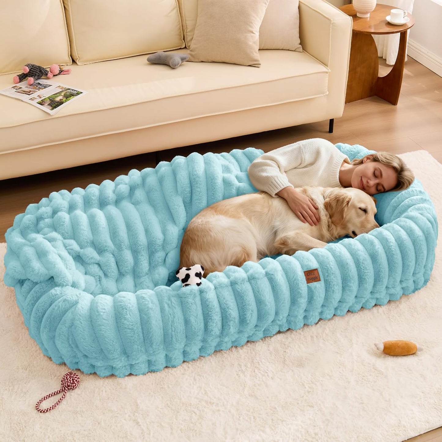 Woman lying on a large blue pet bed with a dog, in a living room setting.