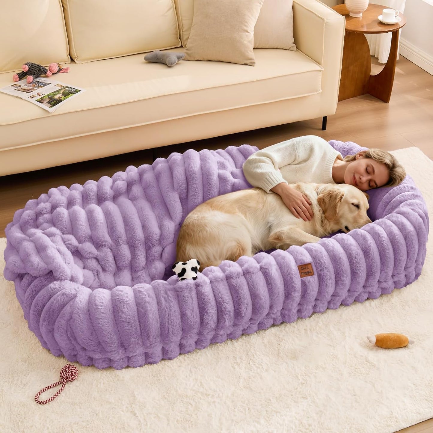 Woman lying on a large purple pet bed with a dog in a living room.